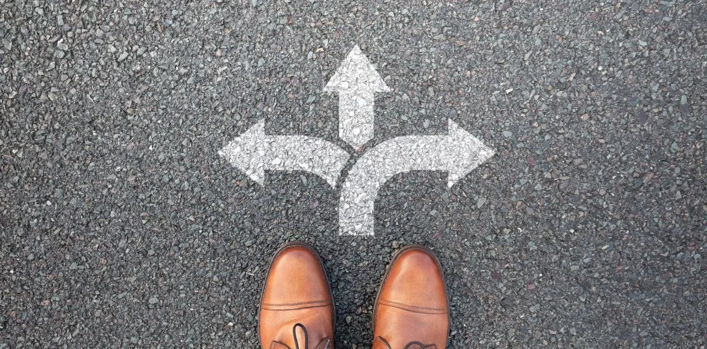 A man stands on a concrete wearing jeans and brown shoes. In front of his feet are 3 arrows going different directions signifying his having a few different choices he can make. In this case, he is choosing between concrete polishing or epoxy coatings.