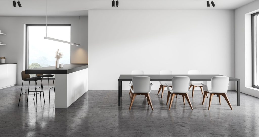 Modern, minimalist home with concrete flooring and white walls. There is a grey dining table and 6 white chairs surrounding it. 2 large windows let lots of natural light into the space. The kitchen has a bar with two stools complimenting the contemporary design. Epoxy concrete flooring is a great modern trend for homes and businesses alike.