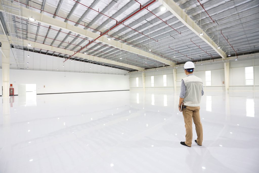 Workman wearing a hardhat and holding a clipboard is standing in the middle of an all white warehouse with white urethane concrete or epoxy coating.