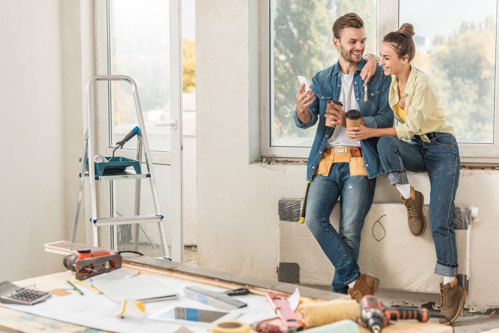 A young couple stands against the wall in their living room which is undergoing home improvements. They are drinking coffee and looking at their mobile phone while making plans. Epoxy flooring is a great home improvement to consider in the spring.