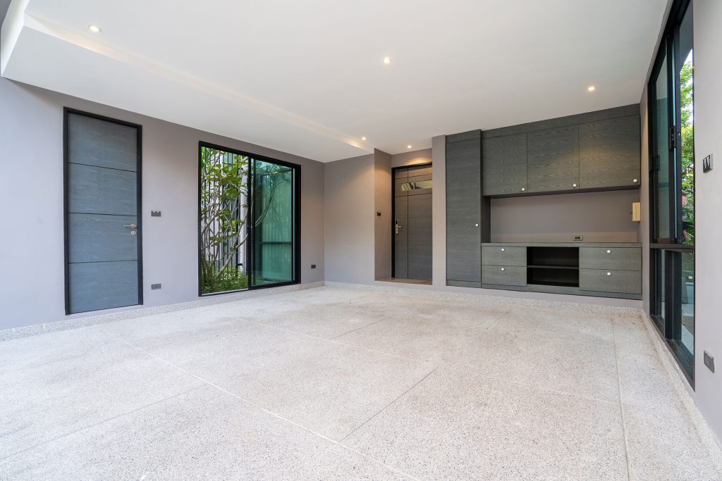 An empty, modern room with light grey walls and large epoxy concrete tiles on the floor and coves on the walls. The space features two dark grey doors, one on the left wall and another on the far wall, along with a sliding glass door leading to an outdoor area with greenery visible through the windows. Built-in dark grey cabinets with silver handles line the far wall, providing ample storage space. Recessed ceiling lights illuminate the room, creating a bright and clean atmosphere.