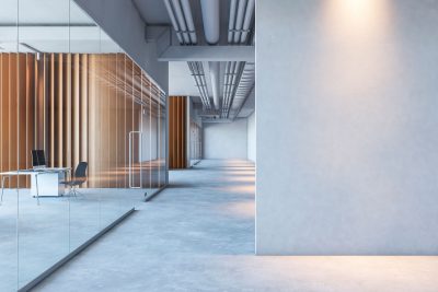 Concrete epoxy floor hallway in an office building, The hall has a modern, industrial feel with concrete walls. Off the hallway are offices with glass doors and wooden walls. In the office sits a desk with a computer, a file cabinet and a chair.