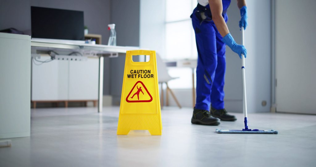 A janitor wearing blue overalls and gloves is mopping the floor in an office environment. In the foreground, a bright yellow "Caution Wet Floor" sign with a warning symbol is prominently displayed on the freshly cleaned floor. A desk with a computer and cleaning supplies can be seen in the background. The overall scene emphasizes the safety and ease of cleaning of epoxy flooring.