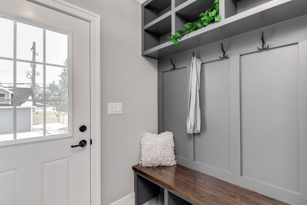 A tidy mudroom with a gray wall featuring built-in hooks, one of which holds a striped cloth. Below the hooks is a wooden bench with storage cubbies underneath, accessorized by a textured white throw pillow. Above the hooks, open cubby shelves are decorated with a small green plant. A white door with large glass panels opens to a snow-covered outdoor scene, adding natural light to the space.