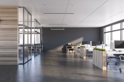 A modern, open office space with sleek, minimalist design. The office features a polished concrete floor, large windows allowing natural light, and several workstations with white desks and ergonomic chairs. Dividers between desks are topped with green potted plants, adding a touch of nature to the space. On the left, there is a glass-walled conference room with a light wood accent wall. The walls are painted in a neutral dark gray, and linear pendant lights hang from the ceiling, contributing to the clean, professional aesthetic.