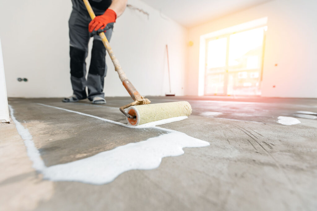 Worker using a paint roller to apply white primer or sealant to a concrete floor in a bright, unfinished room.